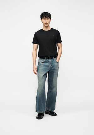 Ung mann iført svart t-skjorte, falmede vide jeans med svart belte, og svarte sko, stående med den ene hånden i lommen.