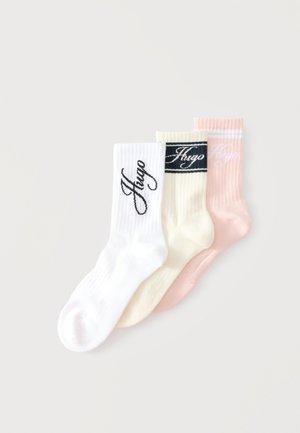 Tre paia di calzini a coste in bianco, crema e rosa, ciascuno con il nome del marchio "Hugo" in diversi stili vicino alla sommità.