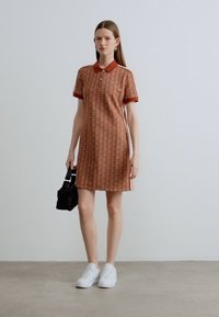 Terrakotta polo ruha geometriai mintával, rövid ujjú, piros gallérral és oldalsávokkal, fehér sportcipővel és fekete táskával párosítva.