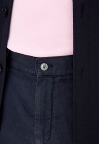Pantaloni in lino blu navy con una finitura texturizzata, dotati di chiusura con bottone e tasche laterali. Sotto si intravede un top leggero di colore rosa chiaro e testurizzato.