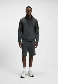 Mörkgrå sportjacka med matchande shorts; lättvikts, textilstruktur; dragkedja, ståkrage och sidofickor. Vita sneakers och strumpor.