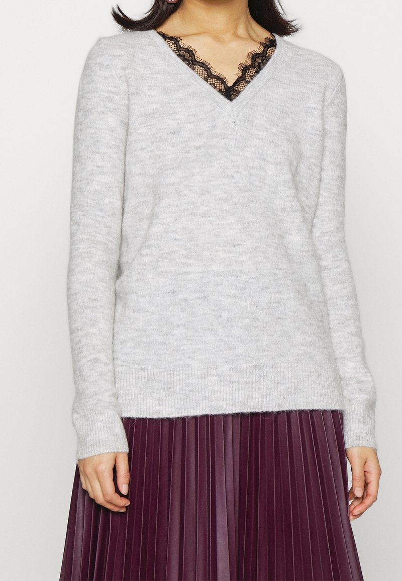 Femme portant un pull gris clair à col en V avec bordure en dentelle noire, associé à une jupe plissée violet foncé, visible du cou aux hanches.