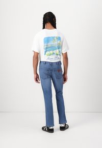 Λευκό γραφικό t-shirt με σχέδιο φοίνικα στην πλάτη, συνδυασμένο με ανοιχτό μπλε τζιν και μαύρες σαγιονάρες.