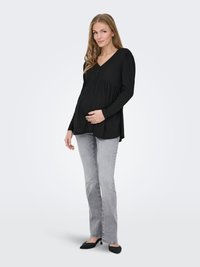 Femme enceinte portant un chemisier noir à manches longues, un jean gris et des talons noirs, debout avec une main sur son ventre contre un fond blanc.