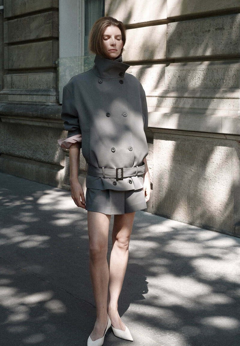 Femme en veste grise oversize à double boutonnage avec ceinture et short assorti, debout sur un trottoir urbain ombragé avec un bâtiment en pierre en arrière-plan.