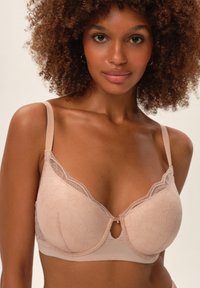 Biustonosz w kolorze cielistym z koronkowym wykończeniem, z fiszbinami i detalem w kształcie dziurki na środku. Gładki materiał oraz regulowane ramiona.