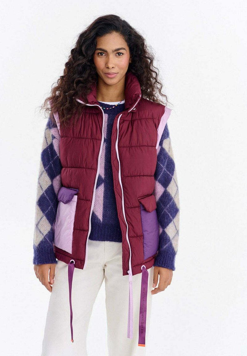 Puchowa bordowa kamizelka z różowymi i fioletowymi akcentami, z przednim zamkiem błyskawicznym, kieszeniami i regulowanymi sznurkami. Nosi się ją na wzorzysty sweter.