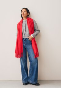 Écharpe en laine rouge à franges drapée sur un cardigan gris, associée à un jean bleu à jambes larges et des chaussures noires. Design simple et texturé.