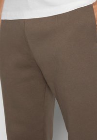 Close-up van een persoon in een bruine, getailleerde broek en een wit shirt, met de focus op het heup- en bovenbeengebied.