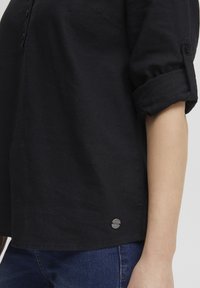 Camicia nera in lino con maniche a risvolto, pattina con bottoni e un accento con logo argentato vicino all'orlo. Tessuto liscio e vestibilità rilassata.