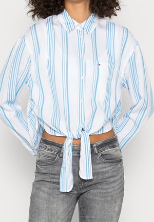 Kobieta w białej koszuli na guziki z pionowymi niebieskimi paskami, zawiązana w talii, w połączeniu z wyblakłymi szarymi dżinsami.
