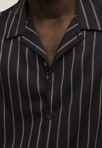 Primo piano di una camicia scura con righe verticali bianche e marroni, dotata di colletto a scannello e bottoni neri, indossata da una persona di pelle scura.