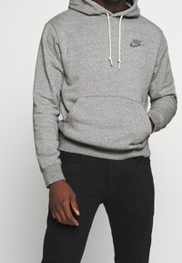 Šedá bavlněná mikina s kapucí, přední kapsou a malým logem Nike na hrudi, v kombinaci s černými kalhotami.