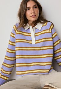Giovane donna con capelli castani che indossa un top a maniche lunghe a strisce lilla, gialle e marroni con colletto bianco e pantaloni beige.