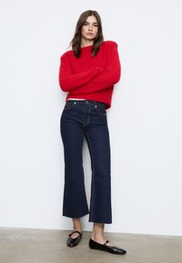 Rdeča teksturirana pletena jopica, temno modri široki kavbojke z kontrastnim šivanjem in črne balerinke. Model stoji s prekrižanimi rokami ob svetli podlagi.