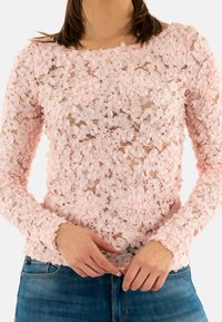 Top in pizzo rosa con applicazioni floreali tridimensionali, maniche lunghe, scollo rotondo e tessuto trasparente. Indossato con jeans in denim blu.