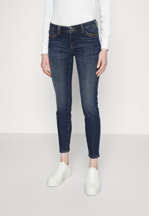 Person iført mørkeblå slim-fit jeans, hvid langærmet top og hvide platform-sneakers står på en ensfarvet lys grå gulv.