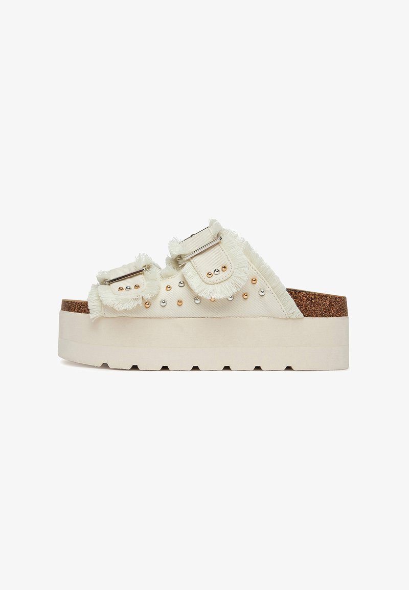Sandalias de plataforma en tela beige claro con detalles de flecos, tachuelas plateadas y doradas, y suela de corcho texturizada. Doble correa con hebillas metálicas.