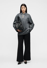 Femme portant un haut à manches longues gris foncé avec un décolleté en forme de goutte, pantalon noir à jambes larges, escarpins noirs pointus, tenant un sac à main en cuir noir.