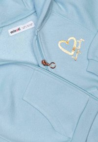 Felpa azzurra con design a cuore dorato, caratterizzata da una chiusura con zip sul davanti e polsini a costine. L'etichetta riporta la scritta "minoti," che indica il marchio.