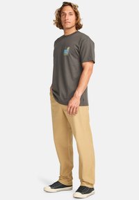 Camiseta de algodón gris con un gráfico colorido en el pecho, combinada con pantalones beige y zapatillas negras. Modelo de pie con las manos relajadas.