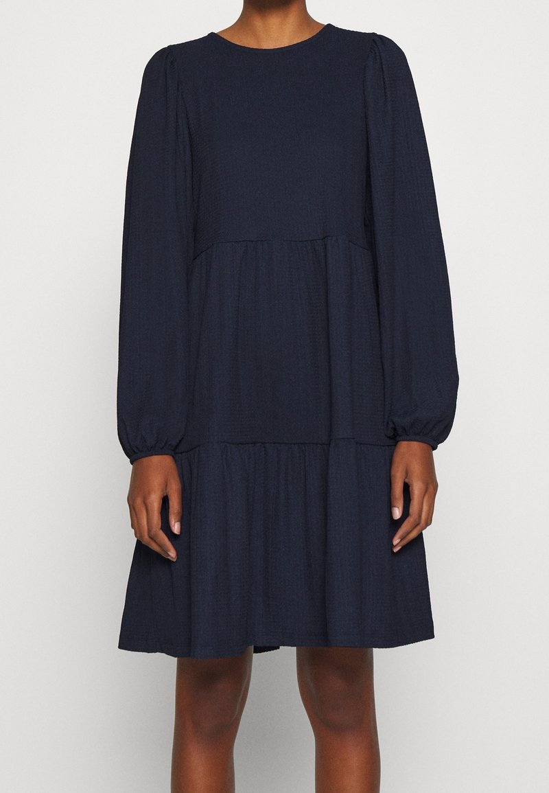 Robe bleu foncé à manches longues, longueur genou, avec une taille froncée et une jupe à volants portée par une personne les mains détendues de chaque côté.
