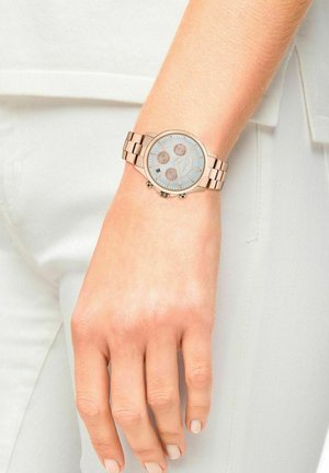 Roségouden chronograaf horloge met een lichtblauwe wijzerplaat, drie sub-dials en een metalen schakelarmband. Hand rust op lichtgekleurde kleding.