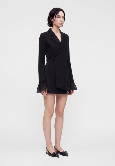 Femme portant une robe blazer noire à manches longues avec des poignets en tulle et des ballerines pointues noires avec un détail de boucle, debout devant un fond uni.