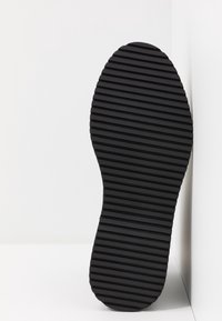 Black rubber shoe sole with horizontal ridged tread pattern, standing upright against a white corner wall and floor.