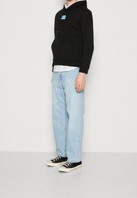 Sudadera negra con capucha y bolsillo frontal, con un logo turquesa, usada sobre una camiseta blanca. Combinada con jeans azul claro y zapatillas negras.