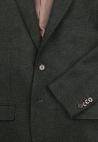 Donkergroene herringbone blazer met een satijnen roze voering, voorzien van een notch lapel, twee voorzakken en vier bruine knopen op de mouwen.