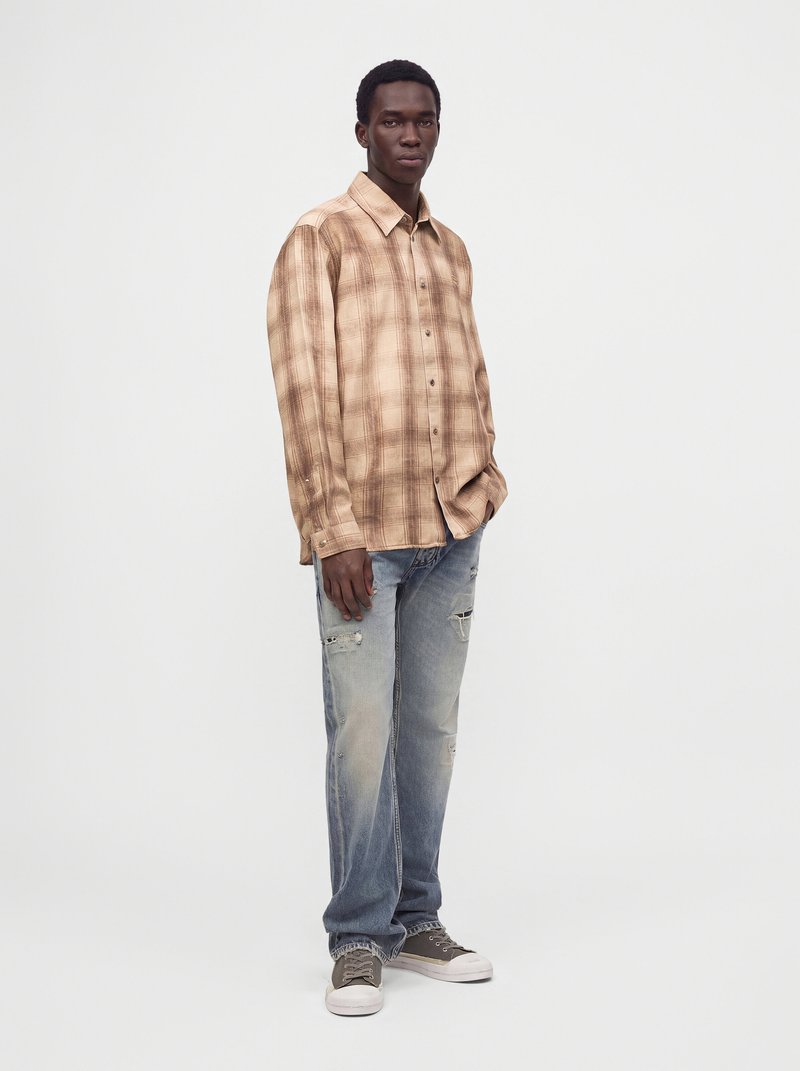 Homme portant une chemise à carreaux marron à boutons, un jean clair délavé déchiré et des baskets grises, debout avec la main dans la poche devant un fond uni.