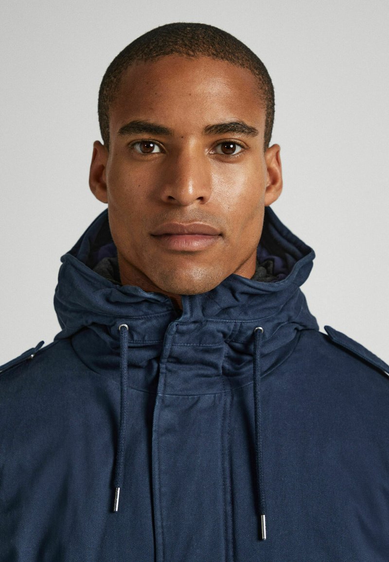 Giacca con cappuccio blu navy dotata di cordoncino, con una texture morbida e bottoni laterali. Il tessuto sembra durevole e leggero.