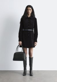 Schwarzes Wildlederhemdkleid mit Druckknopfverschluss, tailliert mit einem Gürtel, kombiniert mit knielangen schwarzen Lederstiefeln und einer strukturierten schwarzen Handtasche.