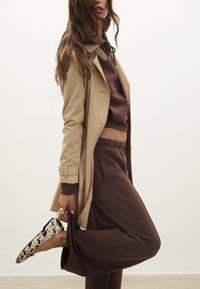 Beige trench coatin päällä ruskea huppari ja vastaavat housut. Mallilla on päällään kerma- ja mustakuosiset tasapohjaiset kengät, ja hän pitää housuista yhdellä kädellä kiinni.