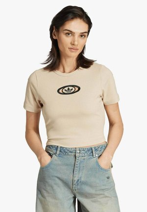 T-shirt beige a maniche corte con righe, logo rotondo nero al centro. Abbinata a pantaloni di jeans azzurri chiari, mani nelle tasche.