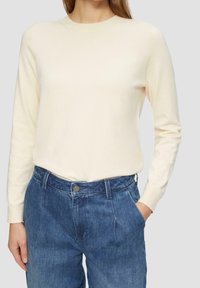 Jasnokremowy sweter z długim rękawem i okrągłym dekoltem, zestawiony z niebieskimi, wysokim stanem spodniami dżinsowymi, z przodem zapinanym na guziki i kieszeniami.