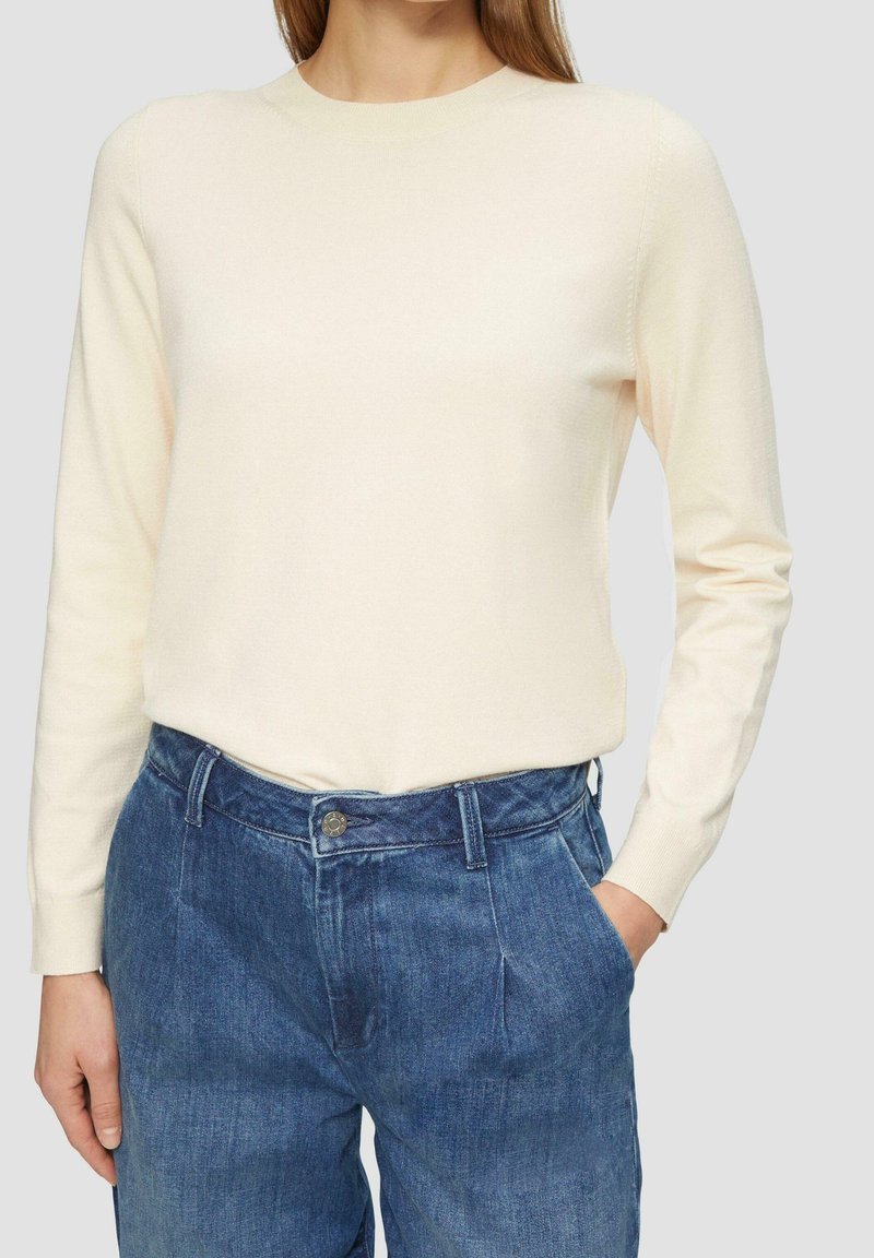 Jasnokremowy sweter z długim rękawem i okrągłym dekoltem, zestawiony z niebieskimi, wysokim stanem spodniami dżinsowymi, z przodem zapinanym na guziki i kieszeniami.