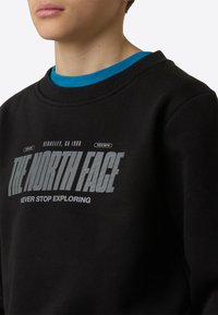 Svart genser med "The North Face"-logo i grått, med teksten "NEVER STOP EXPLORING" under. Ribbet halslinning og ermer.