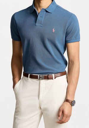 Blaues Polohemd mit strukturiertem Finish, zwei Knöpfen am Kragen und einem rosa Logoakzent. Kombiniert mit beigen Hosen und einem braunen Gürtel.