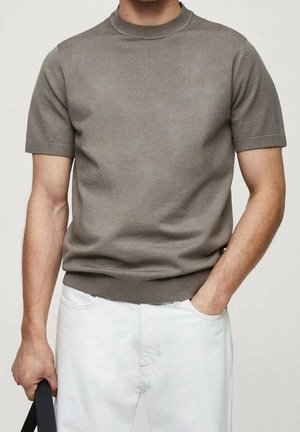 Mann trägt einen kurzärmeligen, grauen Strickpullover mit Rippenkragen und Saum, kombiniert mit weißen Hosen, hält einen schwarzen Riemen in der linken Hand.