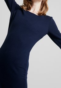 Robe à manches longues bleu marine en tissu texturé avec un motif nid d'abeille, dotée d'un encolure bateau et d'une silhouette ajustée.