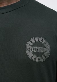 Tamnozelena majica s utisnutim okruglim logom na kojem piše "Versace Jeans Couture" na prsima.