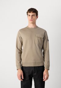 Sweatshirt bege com decote redondo, punhos canelados e um bolso no peito. Confeccionado em tecido macio, combinado com jeans escuros. Design simples e funcional.