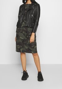 Veste de motard en cuir noir avec des manches côtelées, associée à une robe verte camouflage arrivant aux genoux. Des baskets noires complètent l'ensemble.