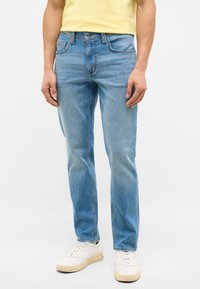 Light blue denim jeans with a straight leg fit, five pockets, and subtle fading. Worn with a yellow shirt and white sneakers.