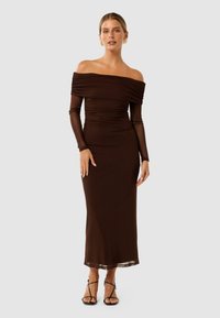 Robe brune décolletée avec des détails froncés, manches longues et une silhouette ajustée, présentant un ourlet transparent et des sandales noires à lanières.