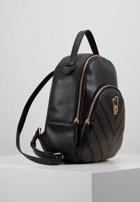 Mochila de cuero negro con cremalleras doradas, que presenta un diseño de chevron con tachuelas en un lado. Incluye un asa en la parte superior y una correa ajustable.
