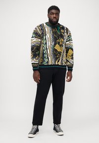 Un pull à motifs noir et jaune avec des accents verts, présentant un design texturé et des poignets côtelés, associé à un pantalon noir et des baskets bleu marine.