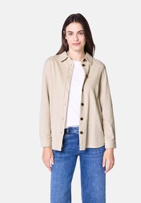 Beige corduroy shirt jacket with a button-up front, pointed collar, and long sleeves; features brown buttons and ribbed texture.